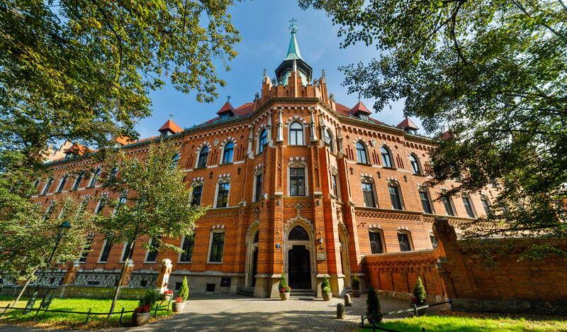 Róg budynku Wyższego Seminarium Duchownego Archidiecezji Krakowskiej. Ceglany trzypiętrowy budynek z wysokimi oknami stoi przed zielonymi plantami.