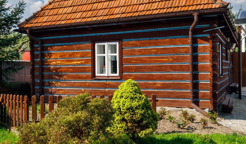Stara drewniana chata w pogodny dzień, będąca Domem bł. Karoliny Kózkówny we wsi Wał-Ruda. Przed chatką znajduje się drewniany płot oraz krzewy.