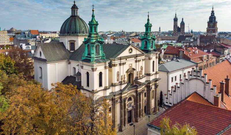 Widok z zewnątrz na Kościół św. Anny w Krakowie. Kościół jest lekko przysłonięty przez drzewo, natomiast widać jego front, dwie wieże i kopułę. Obok widać dachy pozostałych budynków, a nawet Kościół Mariacki i wieżę ratuszową.