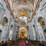 Widok na nawę główną Kościoła św. Anny w Krakowie. Bogato, złoto zdobione stropy, ściany i łuki z malowidłami prowadzą do ołtarza. Na środku kościoła znajdują się też dwa rzędy ławek z krzesłami.