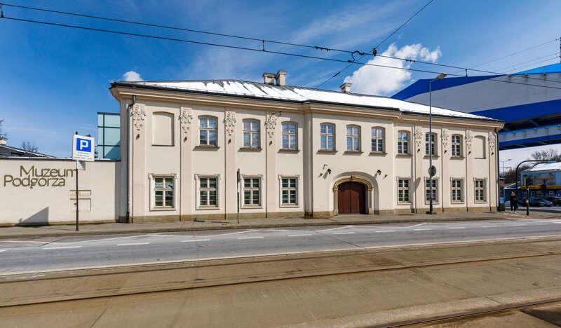 Biały jednopiętrowy budynek Muzeum Podgórza w Krakowie. Przed nim znajduje się ulica z torami tramwajowymi, a nad nimi znajdują się przewody trakcyjne.