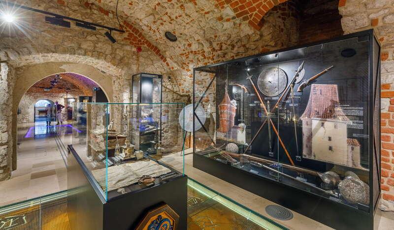 Wnętrze muzeum wyłożone cegłą, wejście do drugiego pomieszczenia zakończone łukiem. Na pierwszym planie gabloty szklane w których są różne eksponaty. Po lewej duża szklana gablota z eksponatami jak miecze.