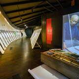 Wnętrze Muzeum Manggha, gdzie po lewej stronie znajdują się drewniane tablice z opisami i zdjęciami. Po prawej duża fotografia mężczyzny przedstawiająca wyrób tkanin. Przed fotografią znajduje się maszyna do  wyrobu przędzy.