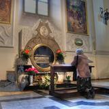 Grób Anieli Salawy w kościele św. Franciszka z Asyżu. Ołtarz boczny z okrągłą wnęką przysłonięta szybą, za nią trumna ze szczątkami błogosławionej Anieli, Nad wnęką okrągły portret.  Na ołtarzu kwiaty, przed nim na klęczniku modląca się starsza kobieta.