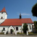 Murowany biały kościół widziany z boku, z zewnątrz. Dach z czerwonej blachy. Od frontu wieża zegarowa.