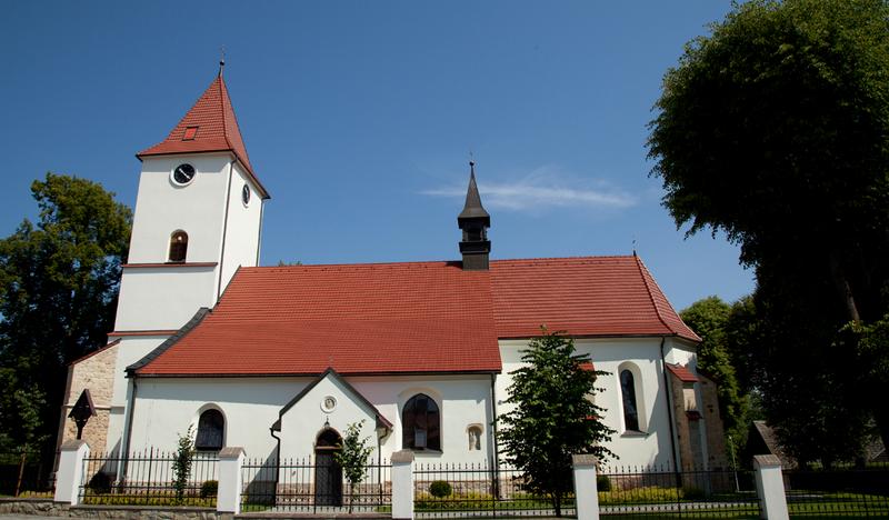 Murowany biały kościół widziany z boku, z zewnątrz. Dach z czerwonej blachy. Od frontu wieża zegarowa.