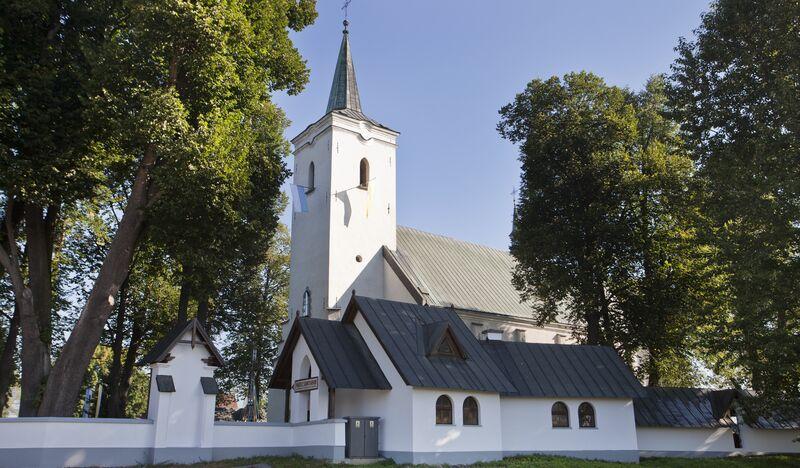 Biały, murowany kościół, pokryty czarnym dachem, z wieżą, murowane białe ogrodzenie.