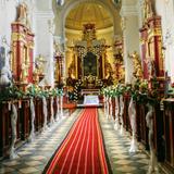 Wnętrze kościoła z widokiem na ołtarz główny, kościelne stalle ozdobione kwiatami. Podłoga w czarno-białą szachownicę częściowo przykryta czerwonym dywanem.