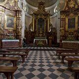 Widok na nawę kościelną z podłogą w czarno-białą szachownicę, po dwóch stronach ławki i ołtarze boczne, na środku bogato zdobiony ołtarz główny.