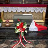 Sarkofag przykryty biało-czerwoną flagą, wokół kwiaty.